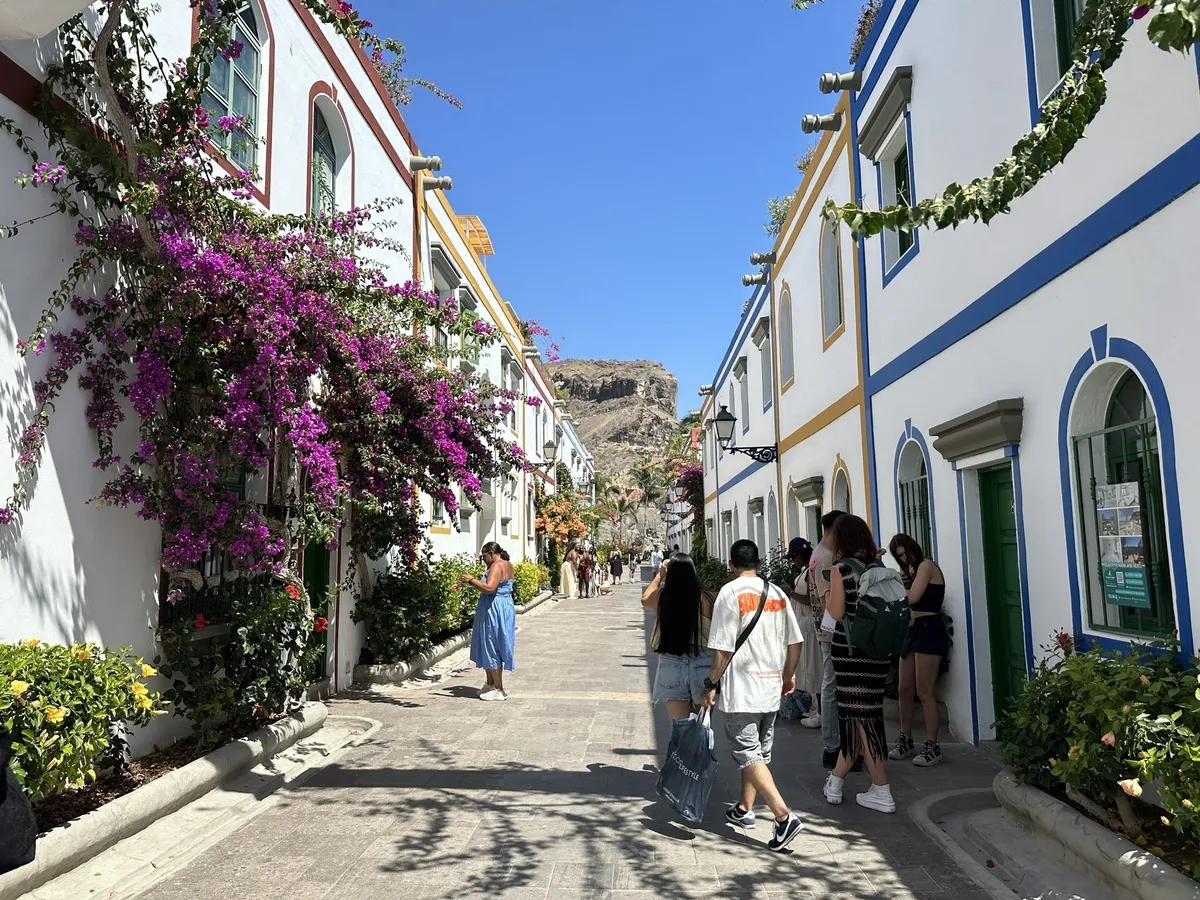 Eine sonnige Straßenszene mit blühenden Bougainvillea-Pflanzen und mehreren Menschen, die zwischen weiß gestrichenen Gebäuden mit farbigen Fensterrahmen spazieren.