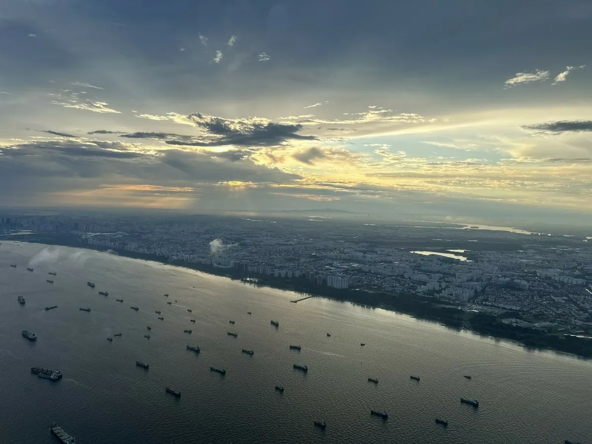 Eine Luftaufnahme bei Sonnenuntergang oder Sonnenaufgang über über Singapur, die sich entlang einer breiten Wasserstraße erstreckt. Zahlreiche große Frachtschiffe ankern auf dem Wasser. Die Stadt liegt unter einem dramatischen, bewölkten Himmel mit durchbrechenden Sonnenstrahlen.