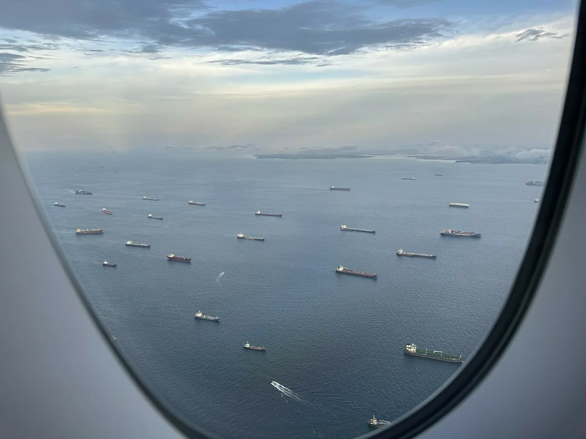 Blick aus einem Flugzeugfenster auf das weite, blaue Meer, das von vielen großen Handelsschiffen befahren wird. Die Schiffe liegen in regelmäßigen Abständen verteilt auf dem Wasser. In der Ferne sind Landmassen und Wolken am Horizont zu sehen.