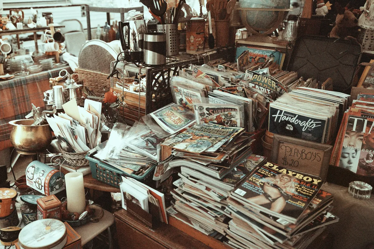 Ein überladener Tisch mit Stapeln von Magazinen und Zeitschriften, darunter Comics und ältere Ausgaben von Playboy. Verschiedene Sammelgegenstände wie Geschirr und Küchenutensilien stehen im Hintergrund.