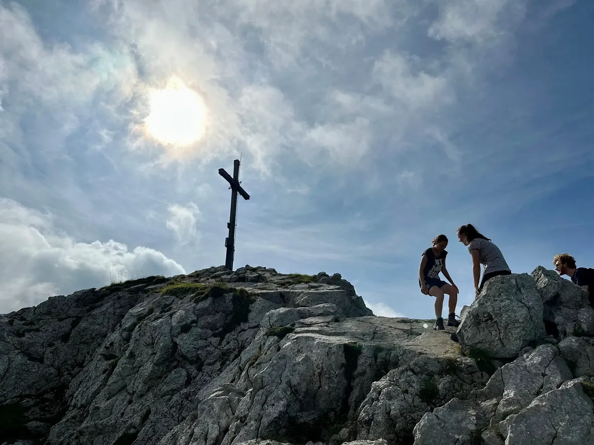 Vier Personen stehen und sitzen auf großen, hellgrauen Felsen an einem Gipfelkreuz unter einem hellen Himmel mit durchscheinender Sonne. Das Holzkreuz ist vor dem strahlenden Licht in Silhouette zu sehen.