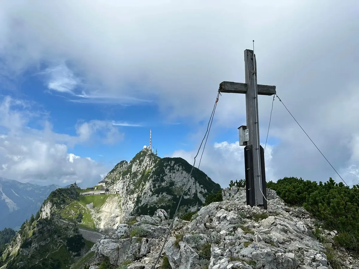 Ein hölzernes Gipfelkreuz auf einem felsigen Gipfel. Im Hintergrund ragt der felsige Gipfel des Wendelsteins mit seinen Gebäuden und Antennen in die Höhe.