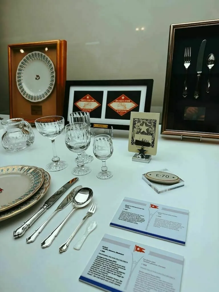 Eine Vitrine mit nachgebildetem Porzellan, Besteck und Glaswaren der Ersten Klasse der White Star Line der Titanic. Im Hintergrund sind gerahmte Exponate und Informationstafeln zu sehen.