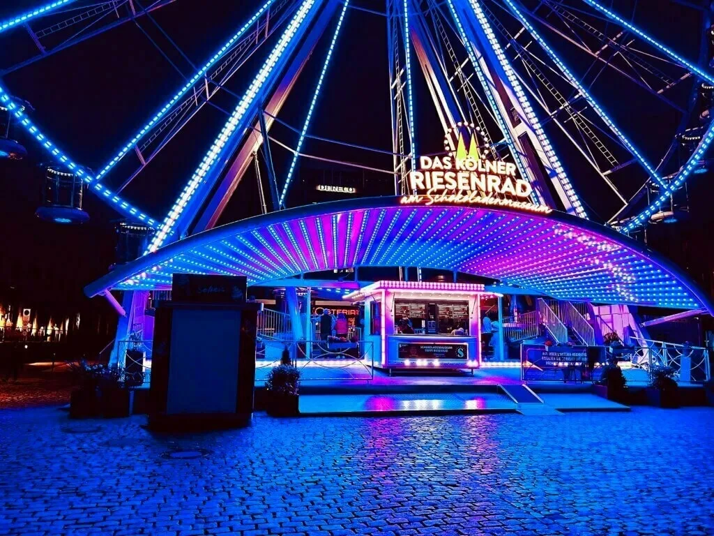 Das Kölner Riesenrad am Schokoladenmuseum bei Nacht, hell erleuchtet in leuchtenden Blau- und Violetttönen.