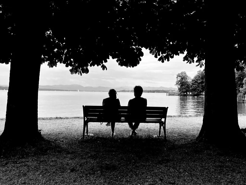 Schwarz-Weiß-Aufnahme, die zwei Personen in Silhouette auf einer Parkbank zwischen zwei großen Bäumen zeigt. Sie blicken auf einen See und eine Bergkette im Hintergrund.