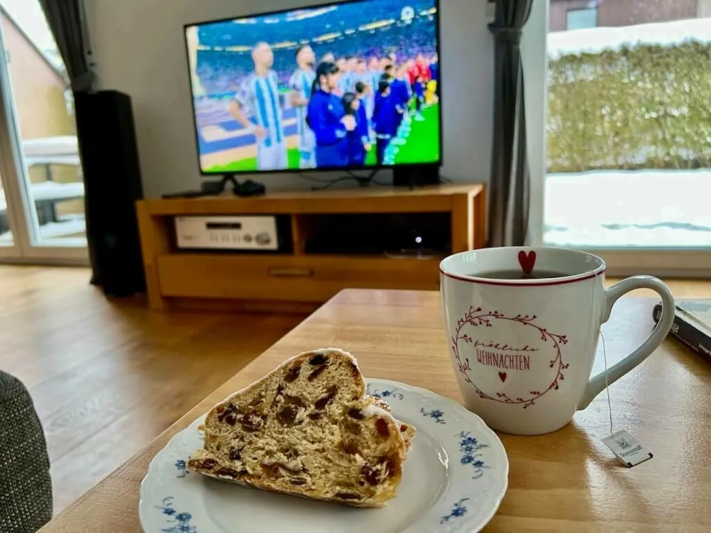 Auf einem Holztisch steht eine Tasse Tee mit einem Stollen auf einem Teller. Im Hintergrund sieht man die argentinische Mannschaft im Fußball-Weltmeisterschaftsfinale 2022 im Fernsehen.