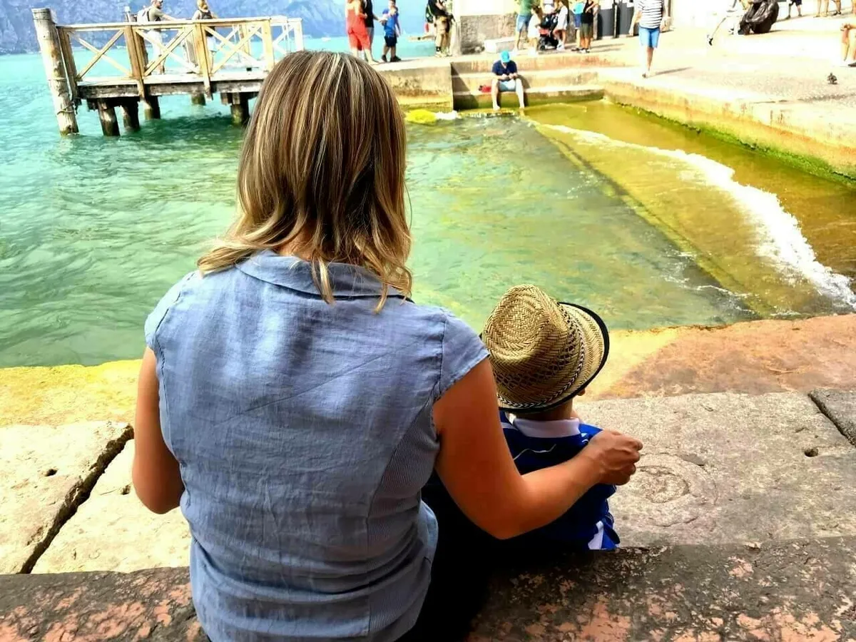 Eine Frau, von hinten gesehen, sitzt mit einem kleinen Kind, das einen Strohhut trägt, am Ufer des Gardasees in Malcesine. Im Hintergrund ist ein hölzerner Steg im Wasser erkennbar.
