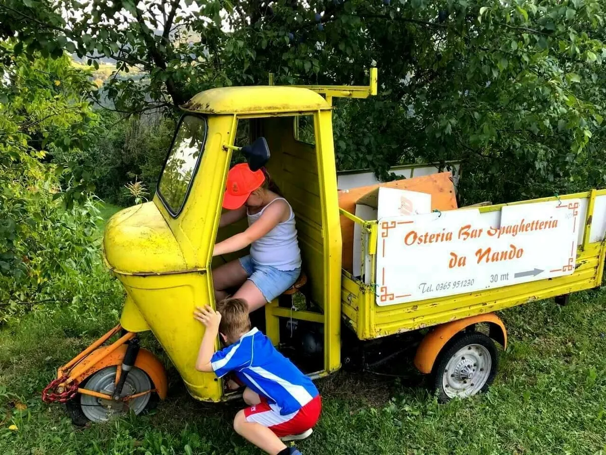 Zwei Kinder spielen in einem gelben, dreirädrigen Kleintransporter, der wie eine Piaggio Ape aussieht. Auf der Ladefläche ist ein Schild mit der Aufschrift "Osteria Bar Spaghetteria da Nando" zu sehen.