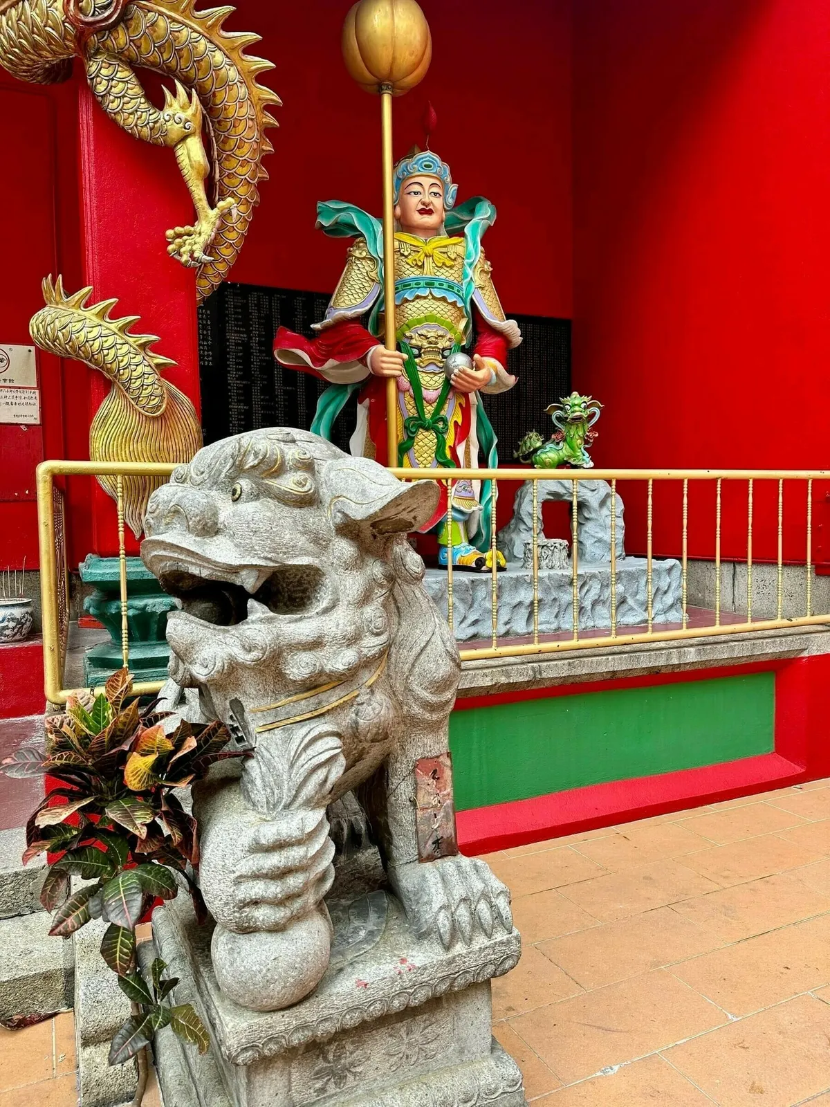 Eine steinerne Wächter-Statue (Foo Dog oder Löwe) im Vordergrund eines chinesischen Tempels. Dahinter eine rote Wand mit zwei goldenen Drachenfiguren und einer farbenfrohen Statue eines chinesischen Gottes oder Wächters in einer Nische.