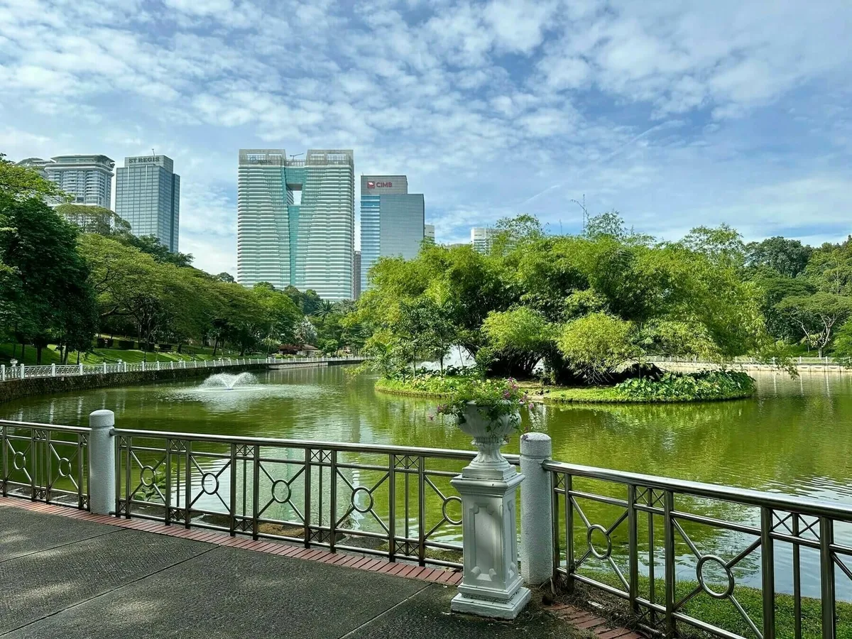 Ein großer Teich in einer Parkanlage mit üppiger Vegetation im Vordergrund und auf einer Insel in der Mitte. Im Hintergrund ragen moderne Hochhäuser in die Höhe. Ein dekoratives Geländer mit weißen Pfeilern verläuft entlang des Uferwegs.