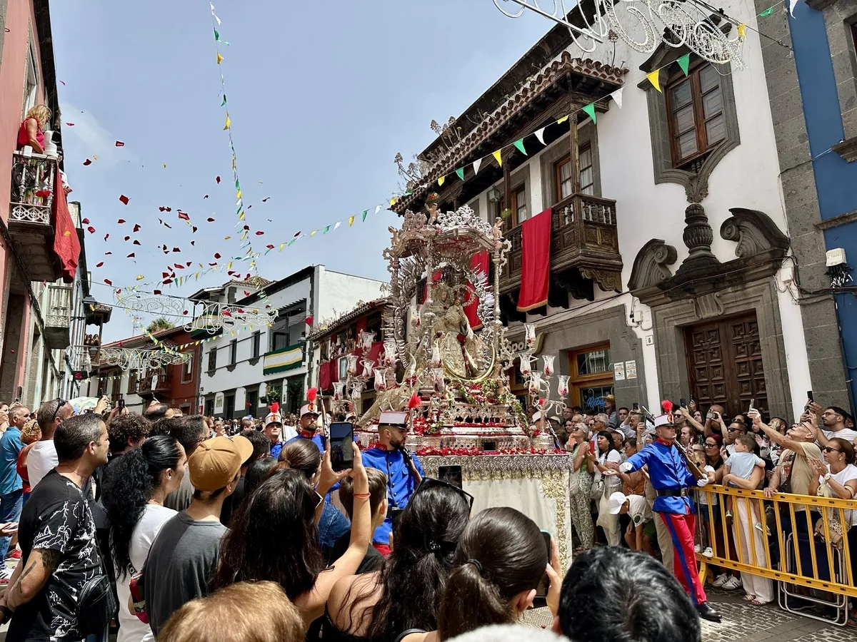 Menschenmengen versammeln sich um den festlichen Umzug zu Ehren der Virgen del Pino in Treror mit einem aufwendig geschmückten Wagen auf einer belebten Straße, während Blumenblätter von oben fallen.