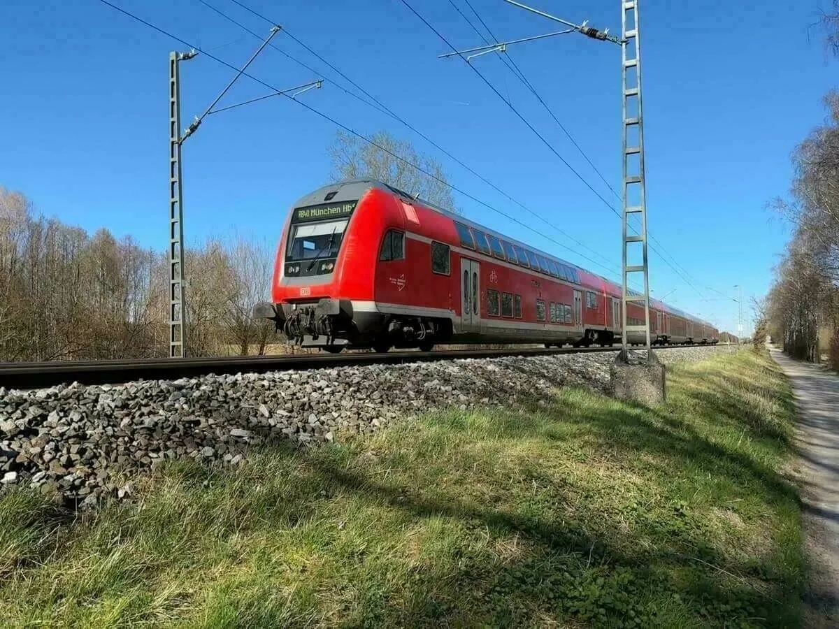 Ein roter doppelstöckiger Regionalzug (DB Regio) fährt bei klarem blauem Himmel auf einer geraden Strecke neben einem Schotterweg.