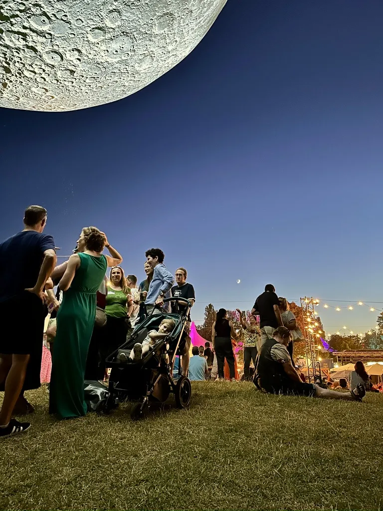 Tollwood 2025 mit Menschen die sich unter einem klaren Abendhimmel unter einem künstlichen Mond versammeln, während festliche Lichter und Zelte die Umgebung beleuchten.