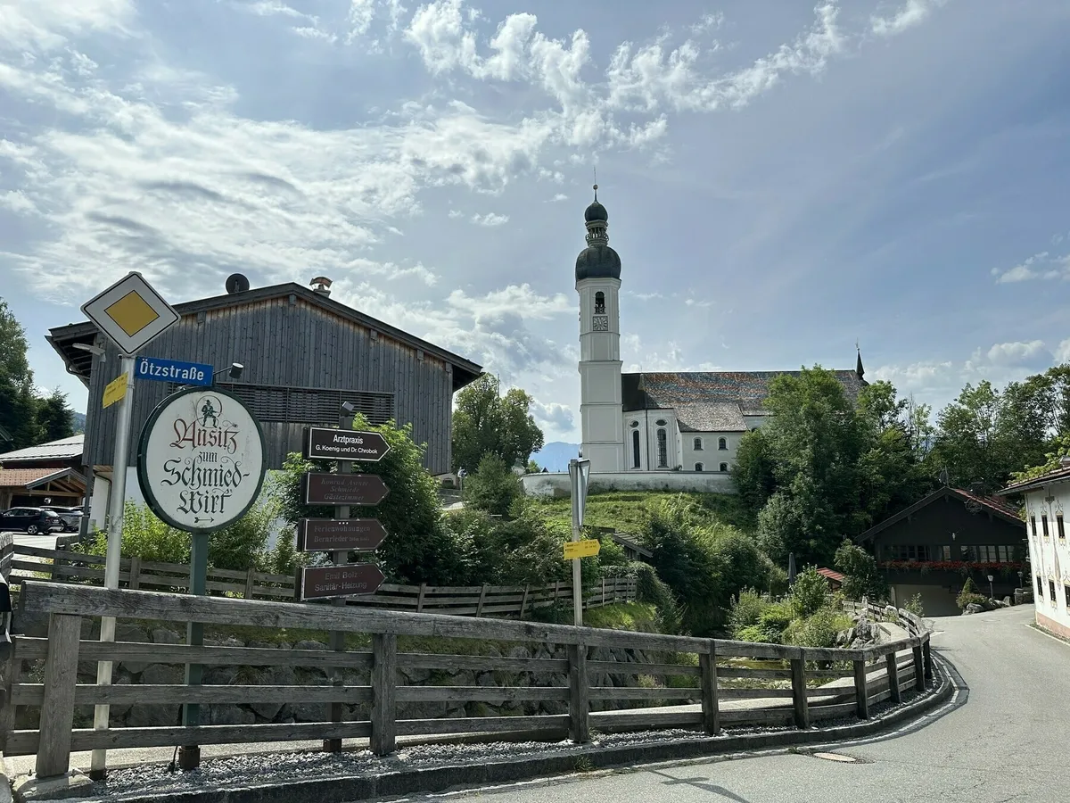 Eine Ansicht einer bayerischen oder alpenländischen Ortschaft. Im Zentrum steht eine weiß verputzte Kirche mit einem hohen Turm und grün-goldener Zwiebelhaube, die auf einem kleinen Hügel liegt. Im Vordergrund sind eine Straße, ein Holzzaun und das Schild des Gasthauses "Zum Schmied-Wirt" zu sehen.