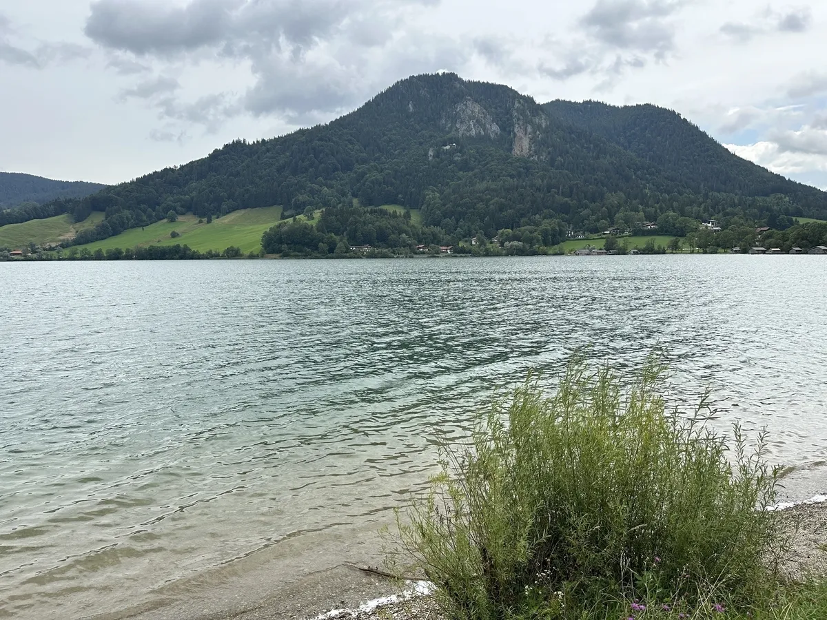 Eine weite Aufnahme eines Sees mit leicht gekräuseltem, hellblau-grünem Wasser. Im Vordergrund wachsen Büsche am Ufer. Dahinter liegt ein großer, dicht bewaldeter Berg mit einigen Häusern an seinem Fuß unter einem bewölkten Himmel.