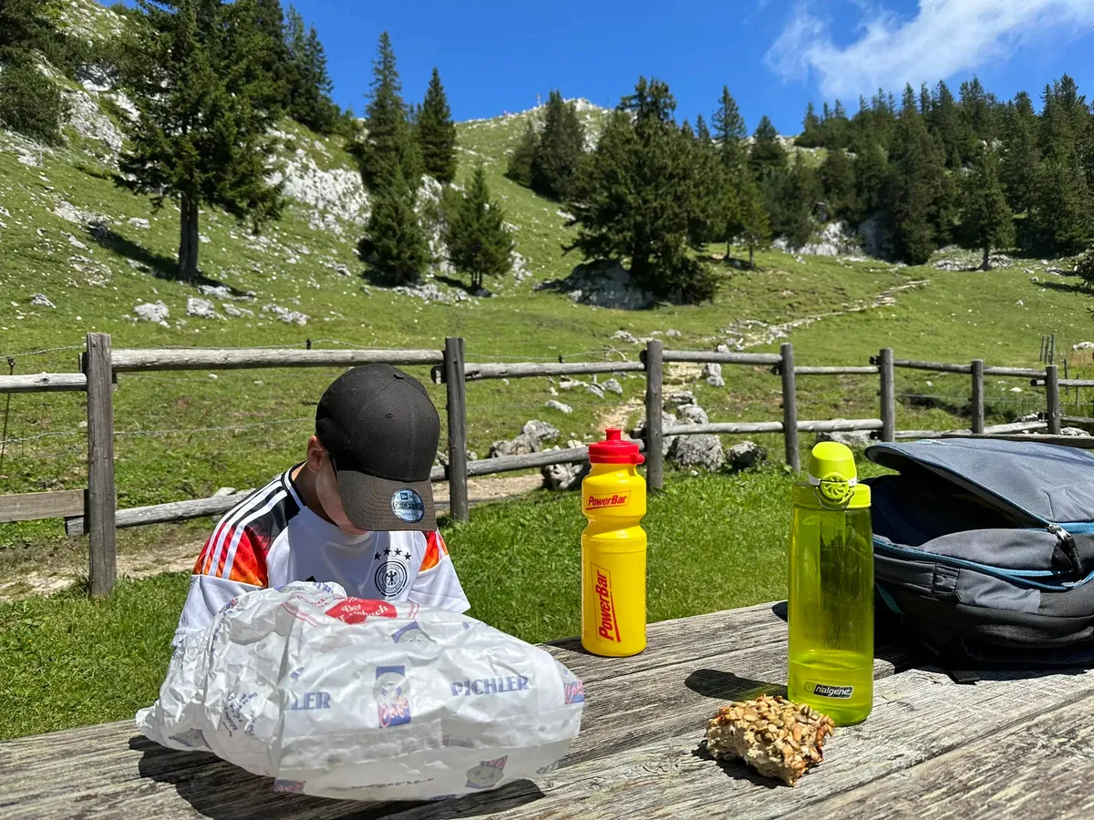 Ein Kind mit einer Kappe und einem Deutschland-Trikot sitzt an einem Holztisch und packt ein eingewickeltes Pausenbrot aus. Neben ihm stehen eine gelbe PowerBar-Trinkflasche und eine grüne Flasche, mit einem Rucksack im Hintergrund.