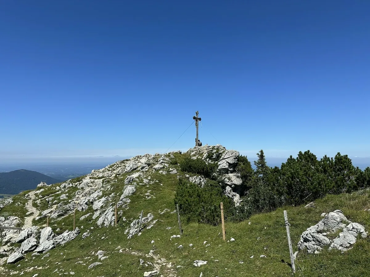 Ein Gipfelkreuz auf einem felsigen, grasbewachsenen Bergrücken unter einem strahlend blauen Himmel. Im Hintergrund ist eine weite Aussicht über die bayerische oder österreichische Voralpenlandschaft zu sehen.