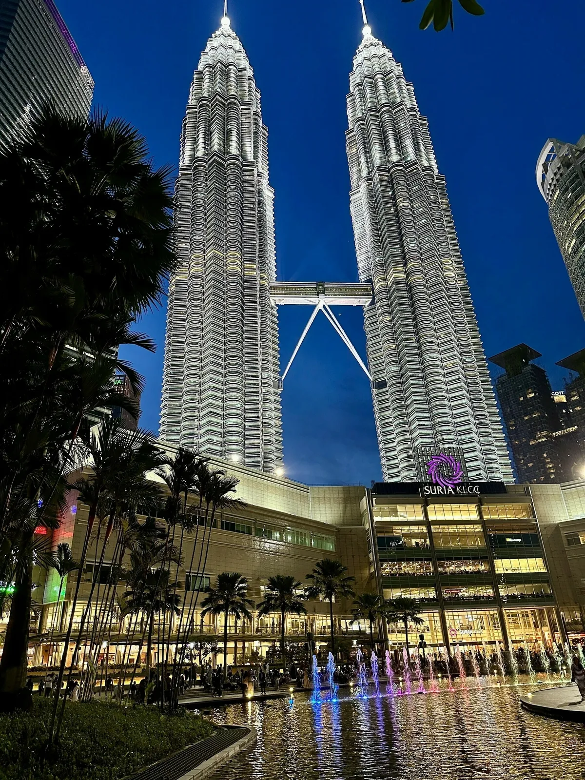 Die Petronas Twin Towers und der davor liegende Suria KLCC Einkaufszentrum bei Einbruch der Dunkelheit. Das Licht des Einkaufszentrums und der beleuchteten Springbrunnen spiegelt sich in einem Wasserbecken im Vordergrund. Palmen umrahmen die Szene vor dem blauen Abendhimmel.