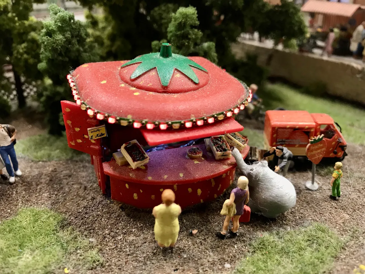 Nahaufnahme eines Miniatur-Erdbeerkiosks auf einer Modellbauanlage. Der rote, erdbeerförmige Verkaufsstand ist beleuchtet und wird von kleinen Figuren sowie einem Miniatur-Ottifanten besucht.