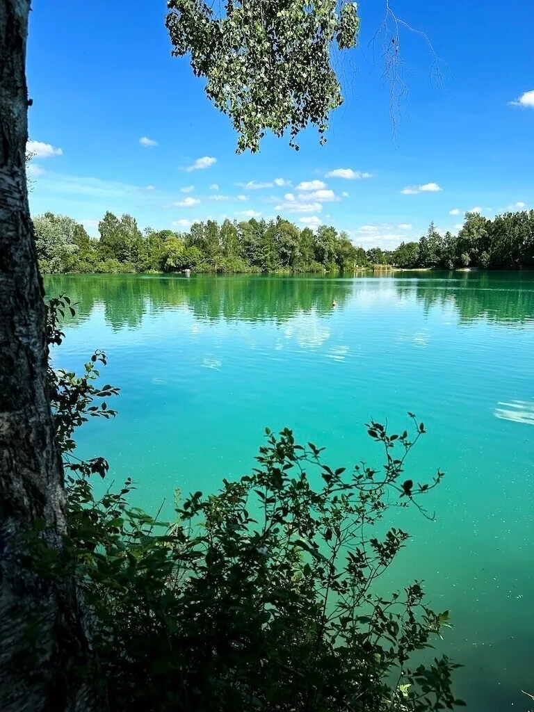 Der klarer, türkisfarbene Wörther Weiher, umgeben von Bäumen und strahlend blauem Himmel.