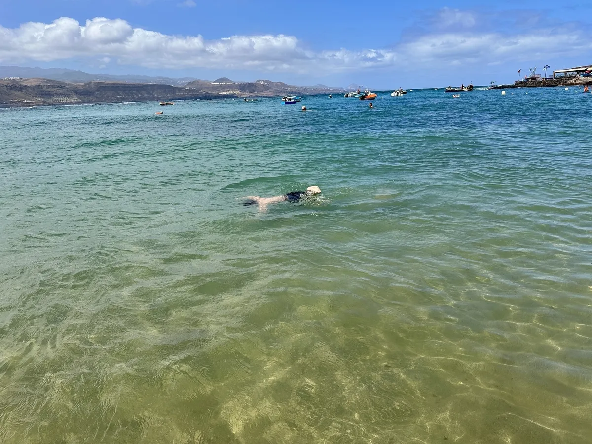 Eine Person taucht im klaren, türkisfarbenen Wasser am Playa de las Canteras, während Boote in der Ferne zu sehen sind.