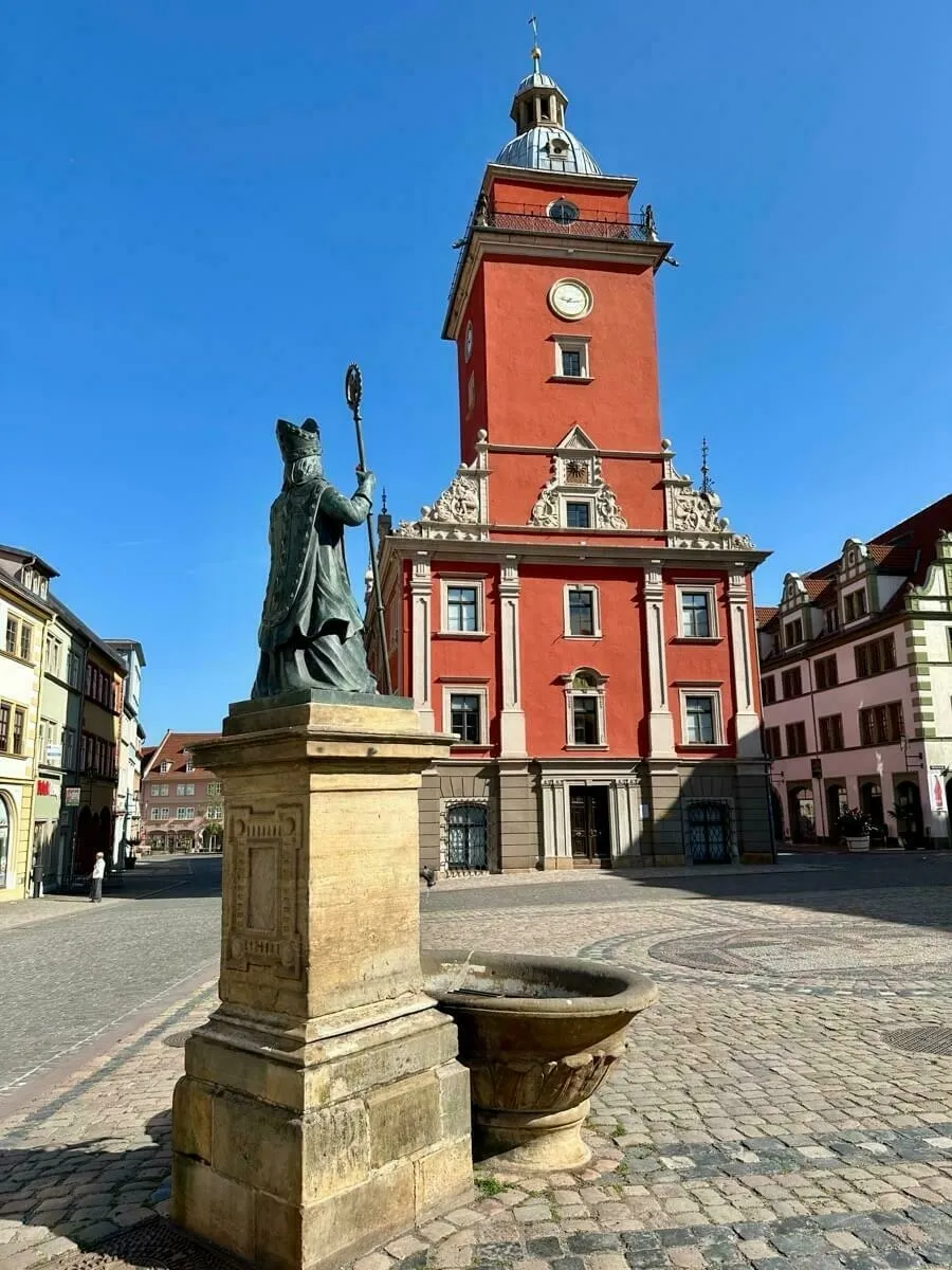 Eine beeindruckende Statue steht vor dem historischen Rathaus mit Uhrturm in einer gepflasterten Platzlandschaft.