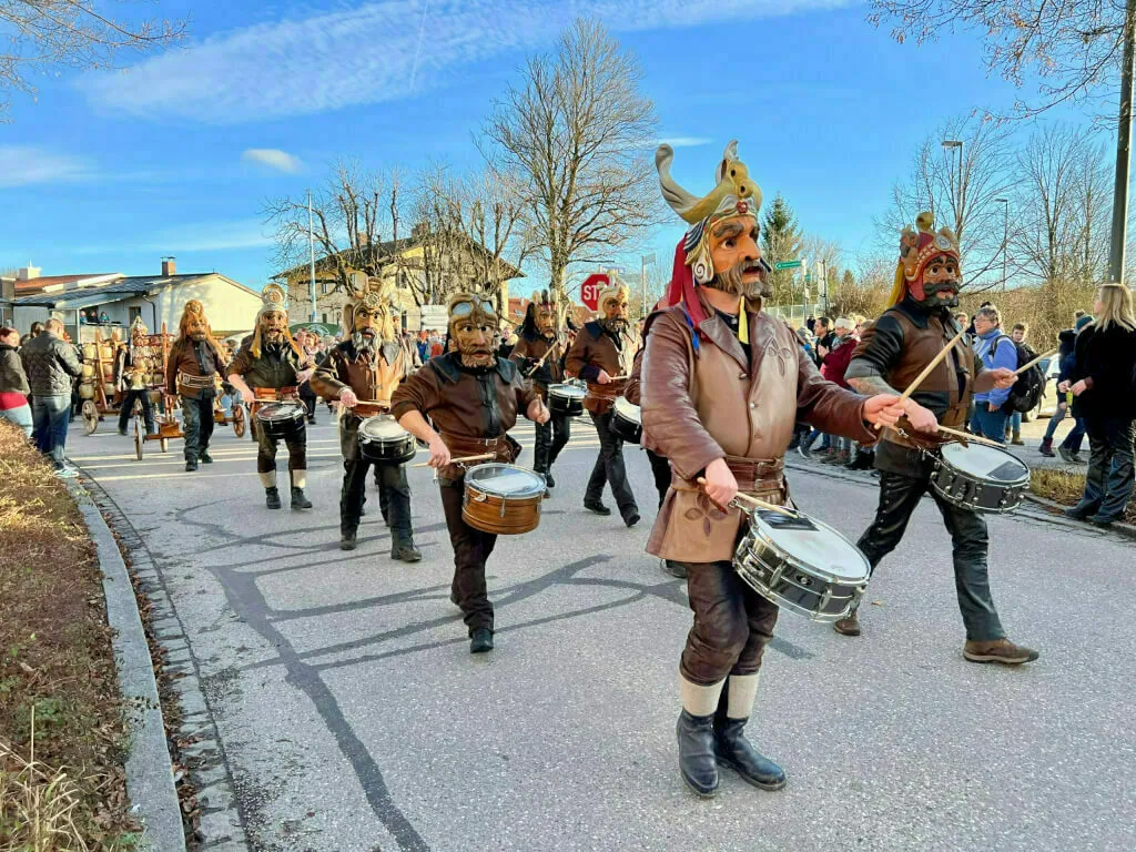 Eine Reihe von Männern in braunen Leder- oder Fellkostümen mit goldverzierten Masken marschiert trommelnd über die Straße.
