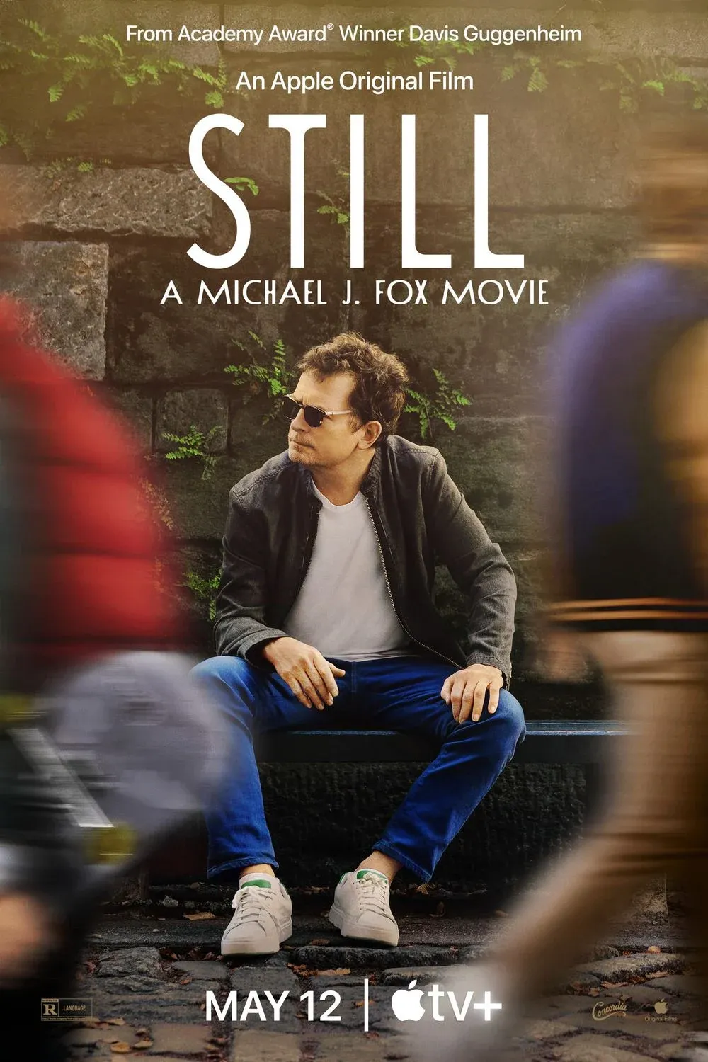 Michael J. Fox sitzt auf einer Bank, während unscharfe Gestalten in Bewegung um ihn herum vorbeigehen, mit dem Text über ihm für einen Film namens Still: A Michael J. Fox Movie.