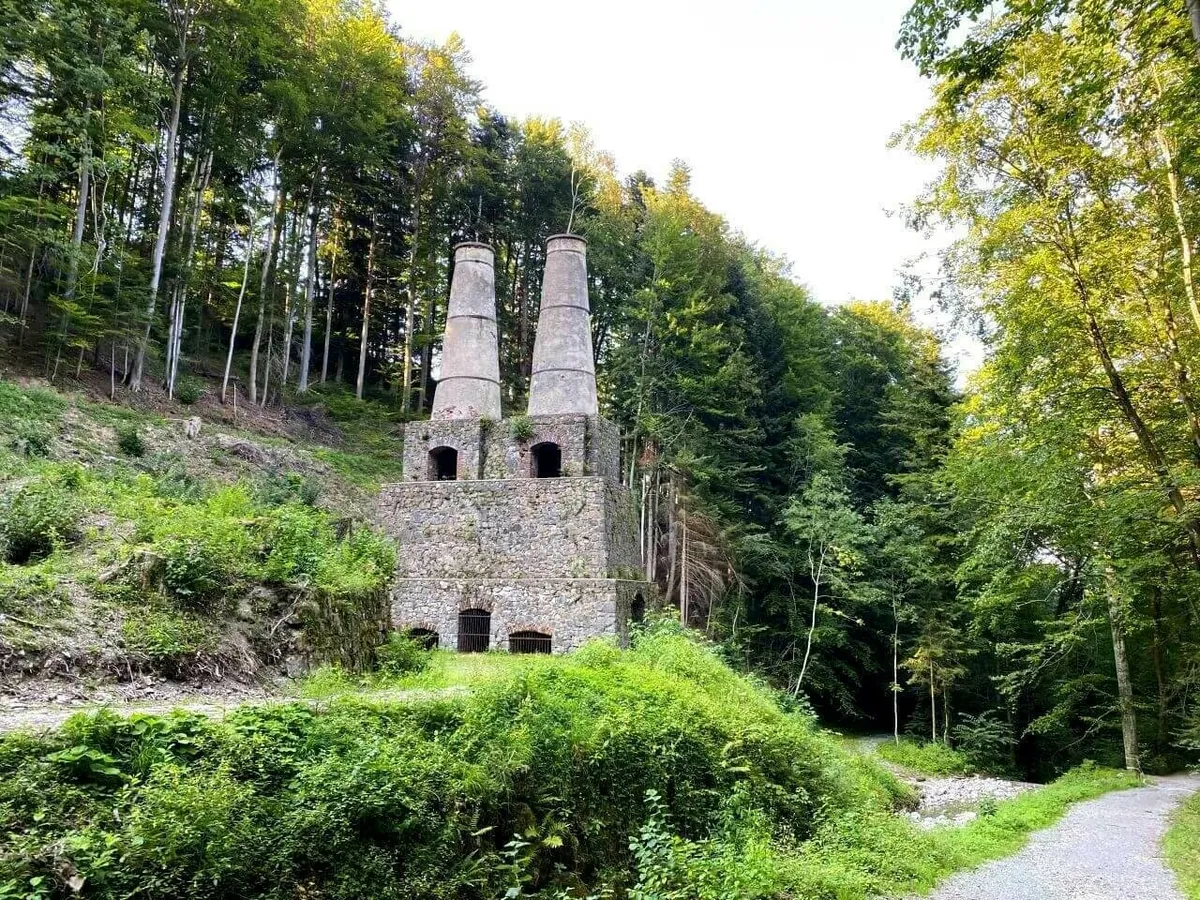 Die steinernen Überreste eines historischen Industriegebäudes, wahrscheinlich des alten Zementwerks in Litzldorf, mit zwei hohen Schornsteinen, umgeben von dichtem Wald.