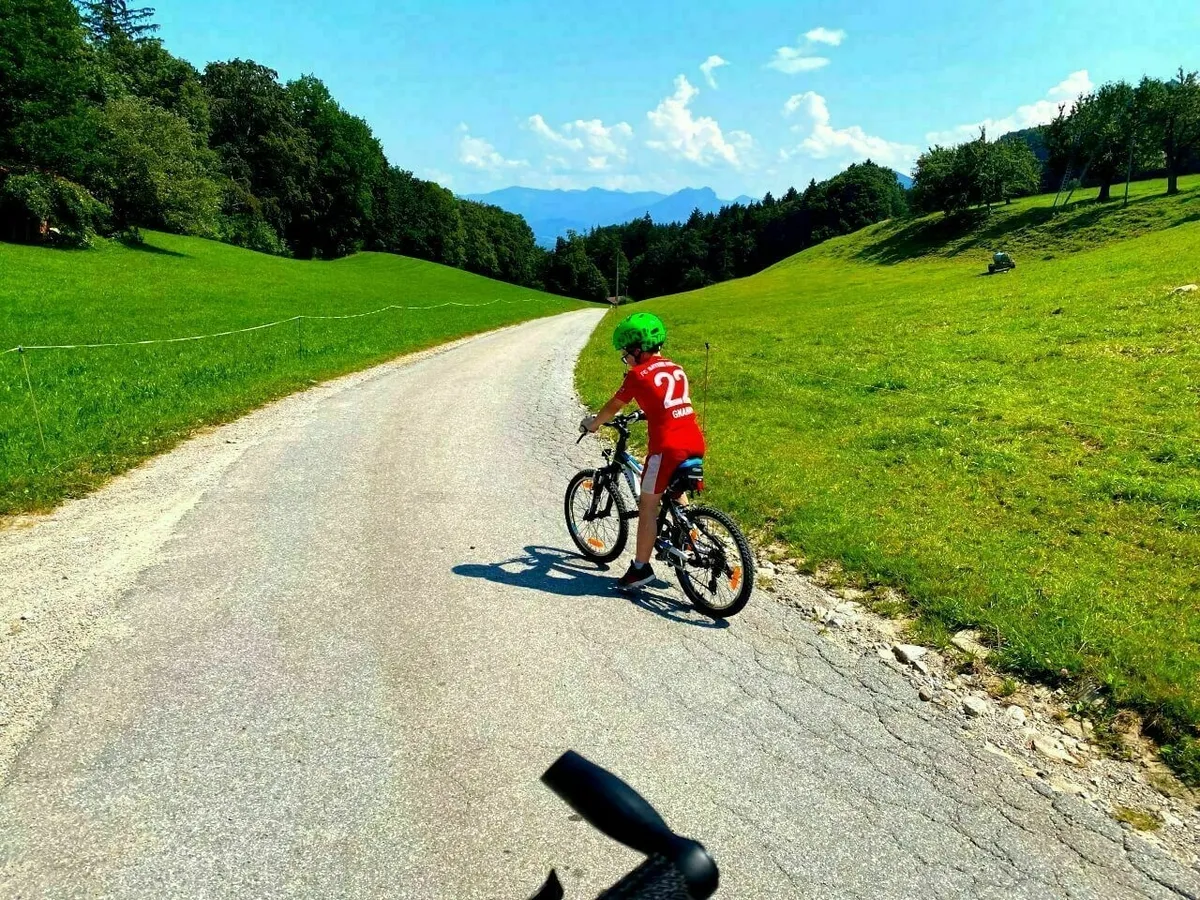 Ein Junge mit rotem Trikot und grünem Helm fährt auf einem Fahrrad eine asphaltierte Straße bergab, die durch grüne Wiesen führt, mit Blick auf Berge in der Ferne.