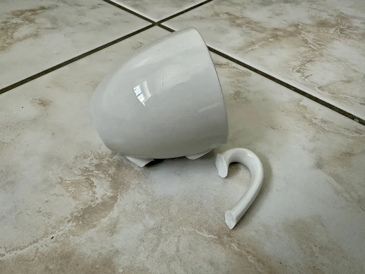 Eine gebrochene weiße Espresso-Tasse liegt auf einem gefliesten Boden. Der Henkel ist abgebrochen und die Tasse ist auf die Seite gefallen.
