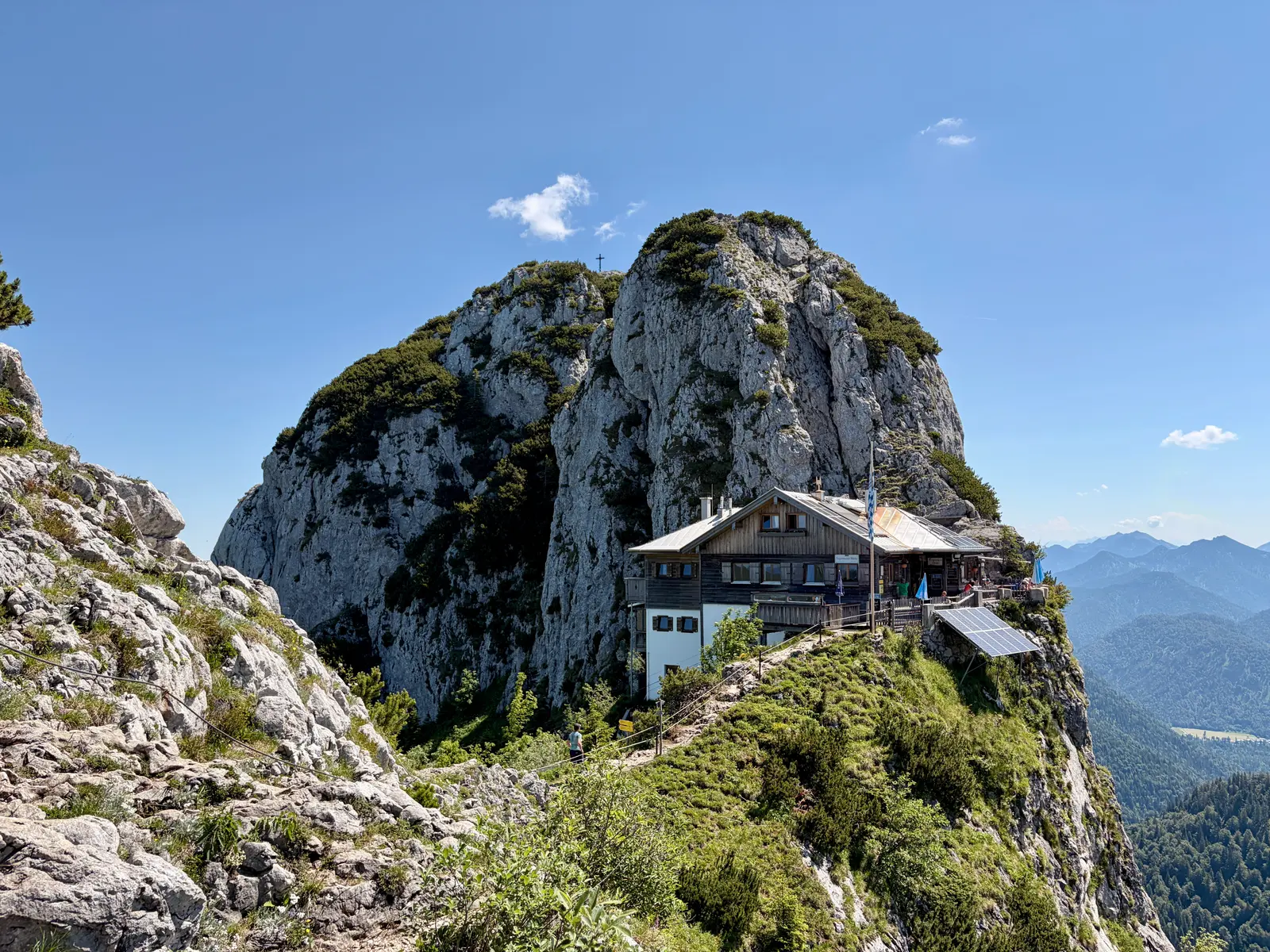 Tegernseer Hütte auf einem schmalen Felsgrat unter dem Buchstein, umgeben von steilen Kalkfelsen und weitem Bergpanorama bei klarem Himmel.