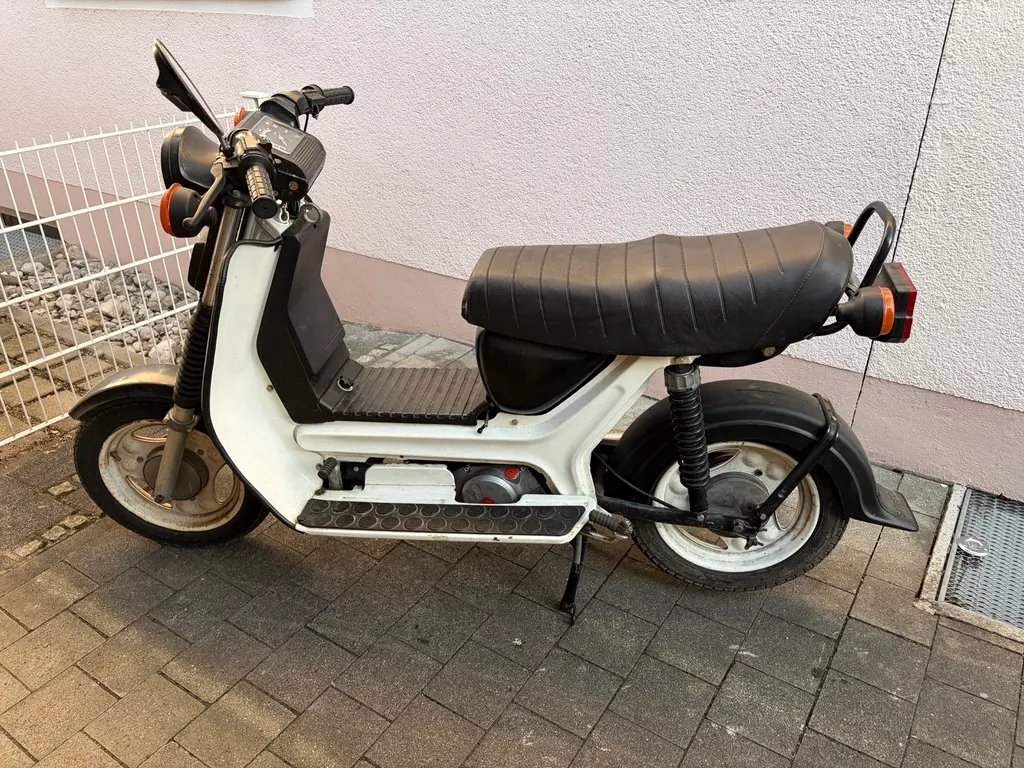 Eine weiß-schwarzes Simson SR50 mit langem schwarzen Sitz steht auf einem Gehweg vor einer hellen Hauswand. Teile wirken abgenutzt, am Lenker und den Rädern sind Gebrauchsspuren zu sehen.