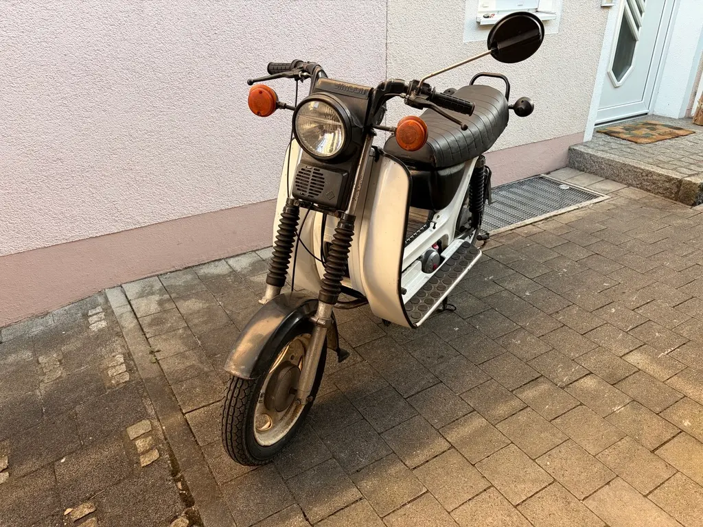 Eine Simson SR50 steht schräg auf einem gepflasterten Gehweg vor einer Hauswand. Vorn sind runder Scheinwerfer, orange Blinker, Spiegel und die beigefarbene Verkleidung gut zu sehen.