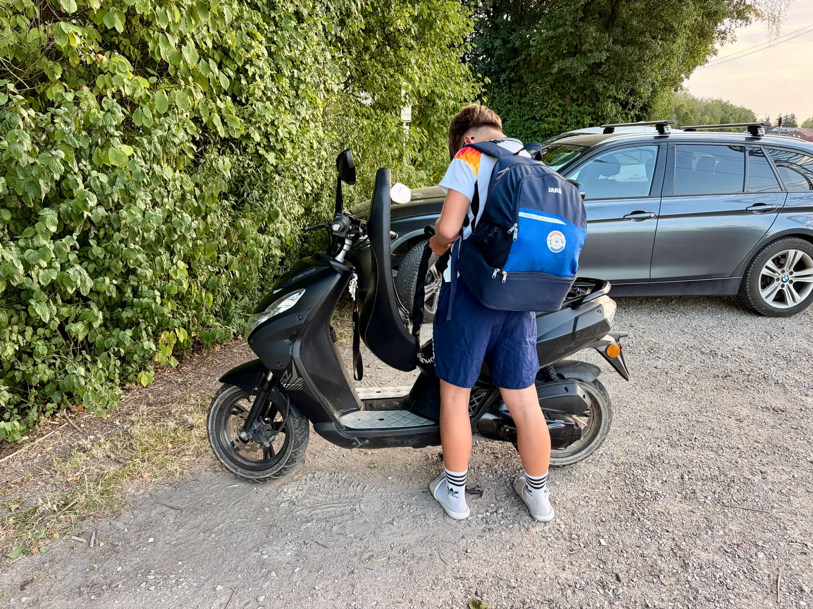 Ein Jugendlicher mit Rucksack steht neben einem schwarzen Roller auf einem Kiesweg und hält einen Helm in der Hand. Im Hintergrund stehen ein Auto und dichtes grünes Gebüsch.