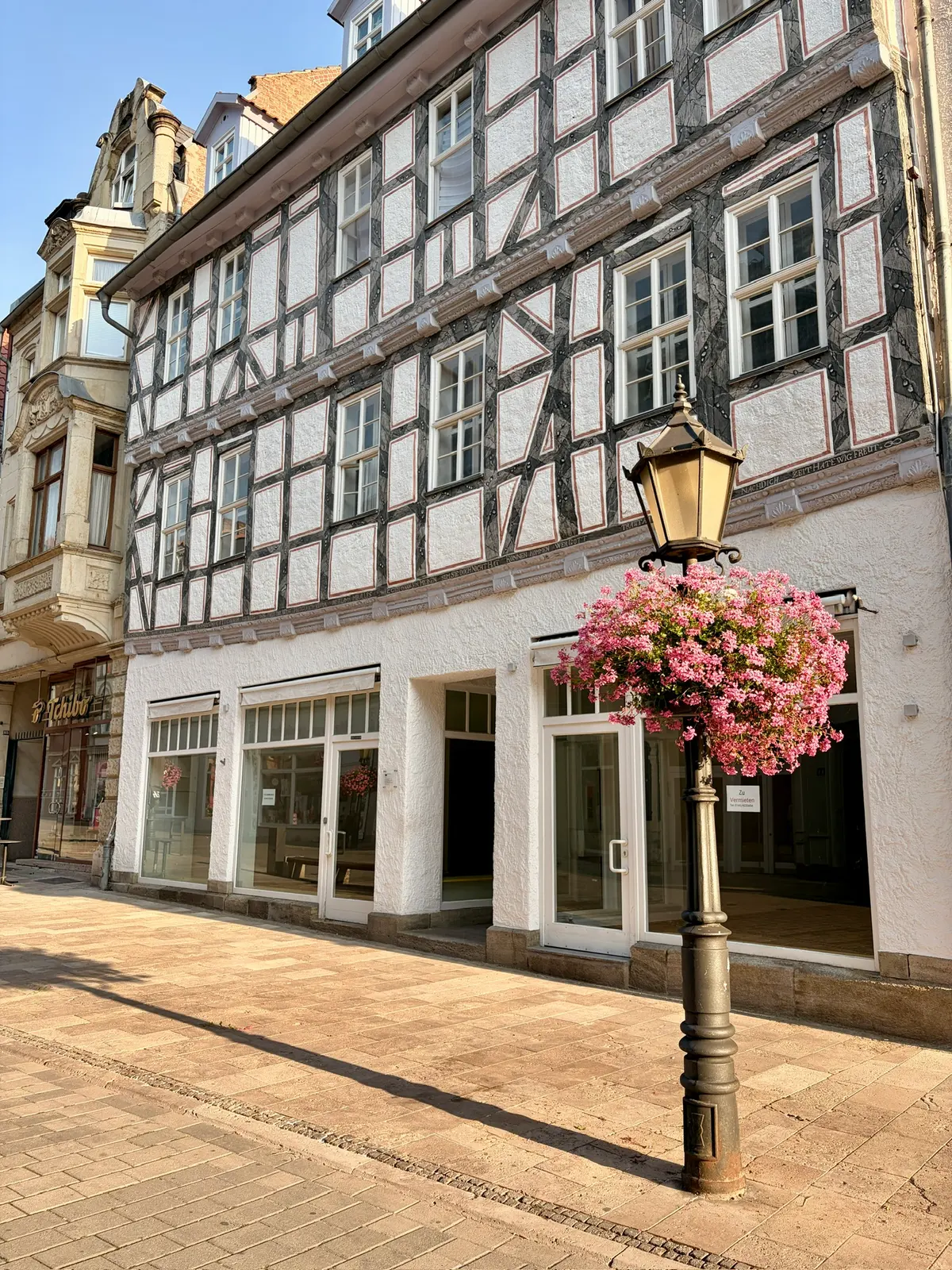 Fachwerkhäuser in einer sonnigen Altstadtgasse, davor eine Laterne mit üppigem rosa Blumenschmuck. Die Ladengeschäfte im Erdgeschoss sind geöffnet, die Straße ist ruhig und menschenleer.