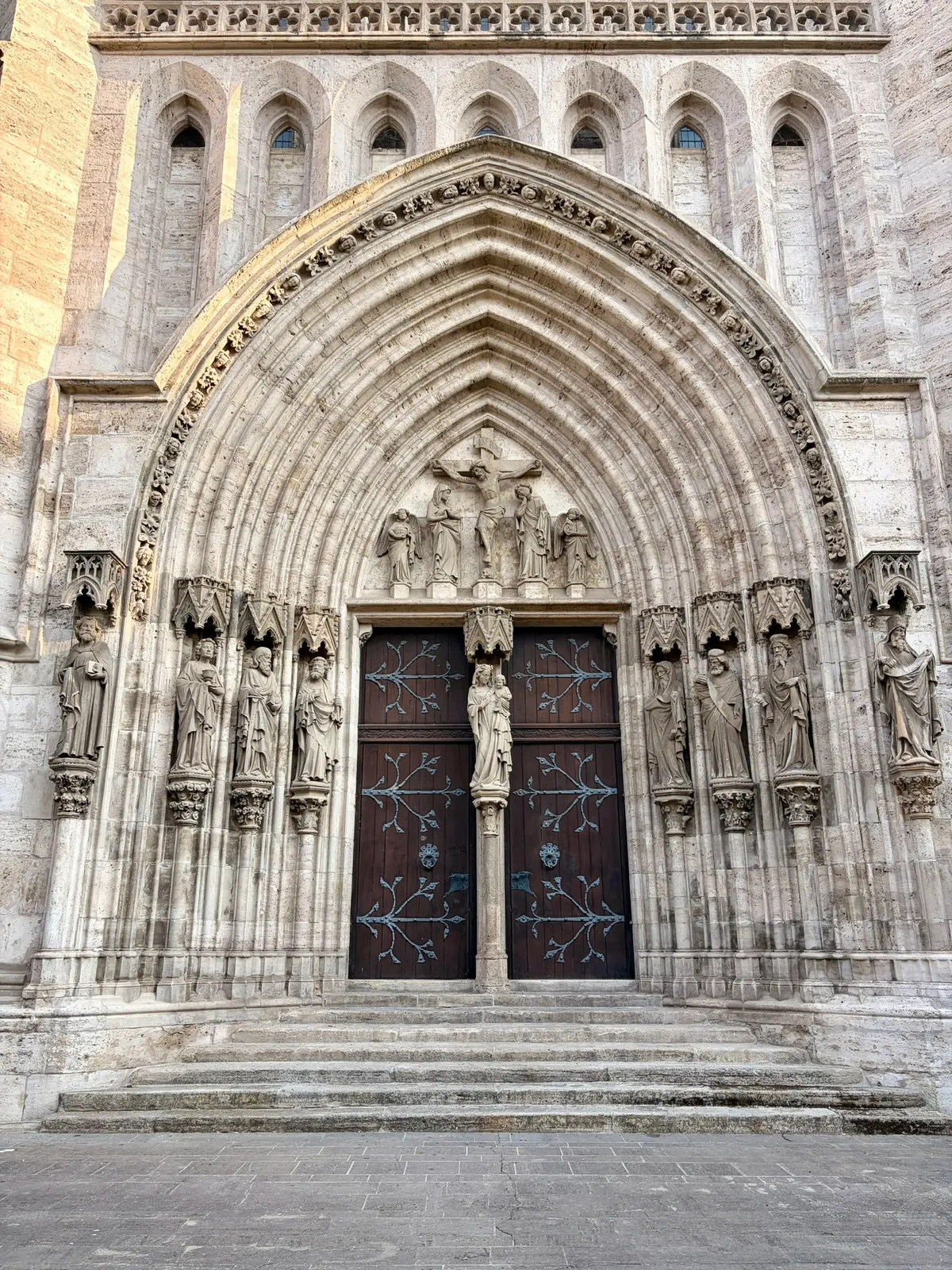 Gotisches Kirchenportal mit reich verzierten Steinbögen, Figuren und einer dunklen zweiflügeligen Tür. Davor führen mehrere breite Stufen zum Eingang hinauf.