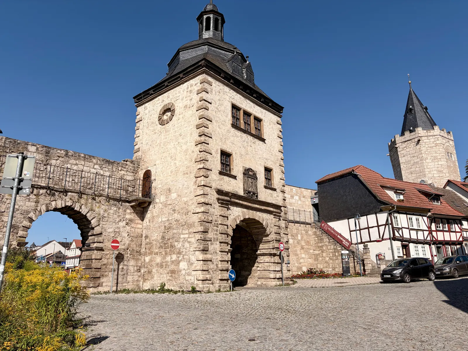 Mittelalterliches Stadttor aus hellem Stein mit Rundbogen und Türmen in Mühlhausen, davor Kopfsteinpflaster und Fachwerkhäuser bei klarem blauem Himmel.