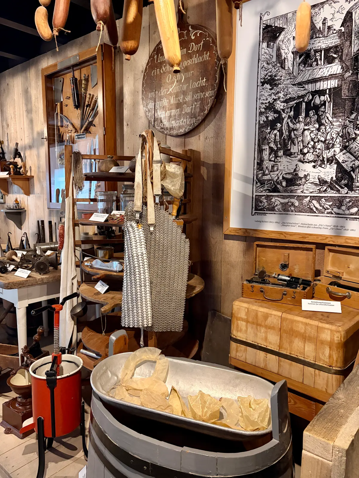 Ausstellung im Bratwurstmuseum mit historischen Geräten, Holzfässern, einer Wurstpresse und einer mit Papier ausgelegten Wanne. An den Wänden hängen Werkzeuge, alte Schilder, hölzerne Wurstmodelle und eine große historische Grafik.
