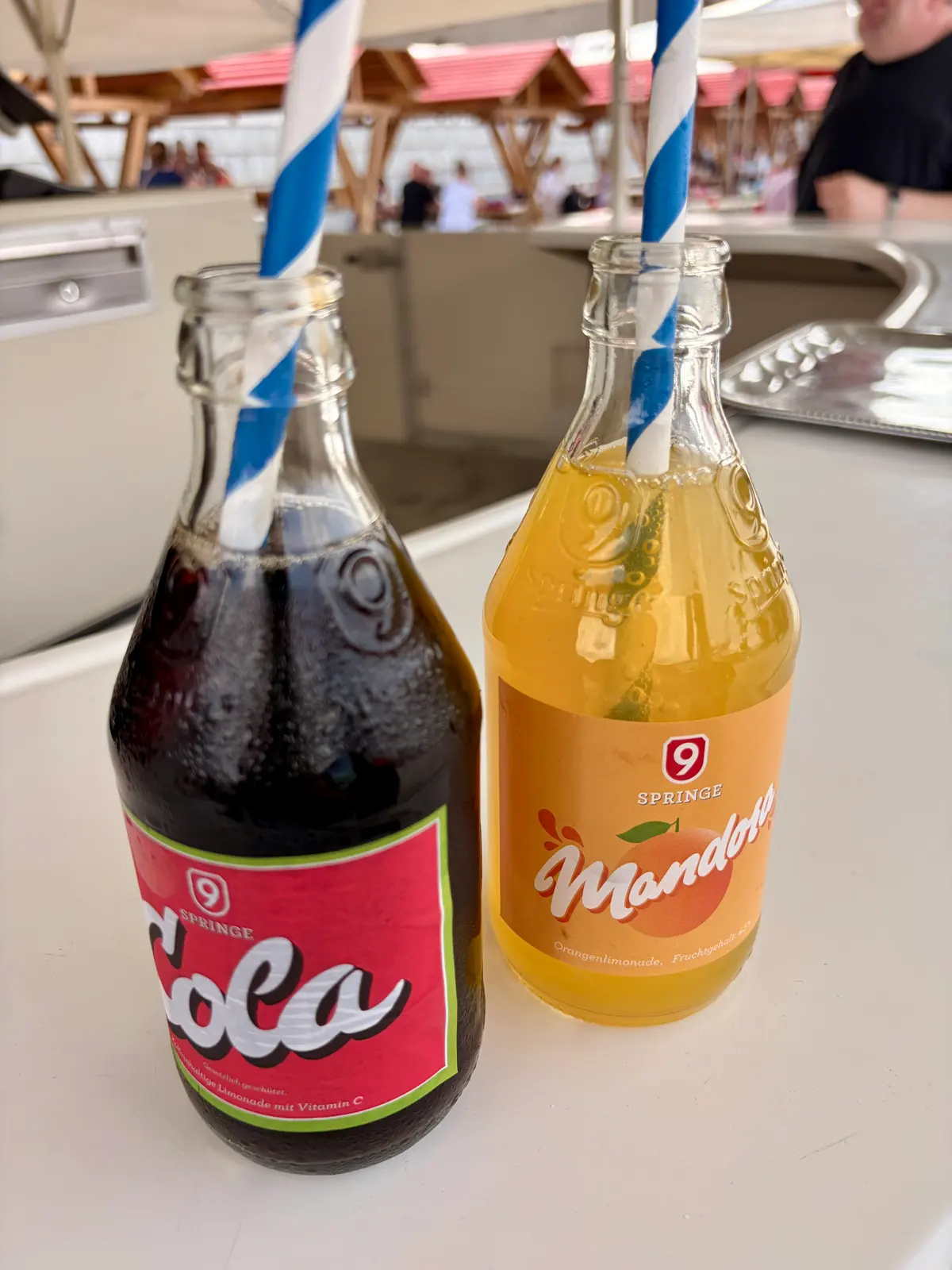 Zwei geöffnete Glasflaschen mit Cola und Orangenlimonade stehen mit blau-weiß gestreiften Strohhalmen auf einem Tisch. Im Hintergrund sind unscharf Sitzplätze und Menschen unter einem Zeltdach zu sehen.